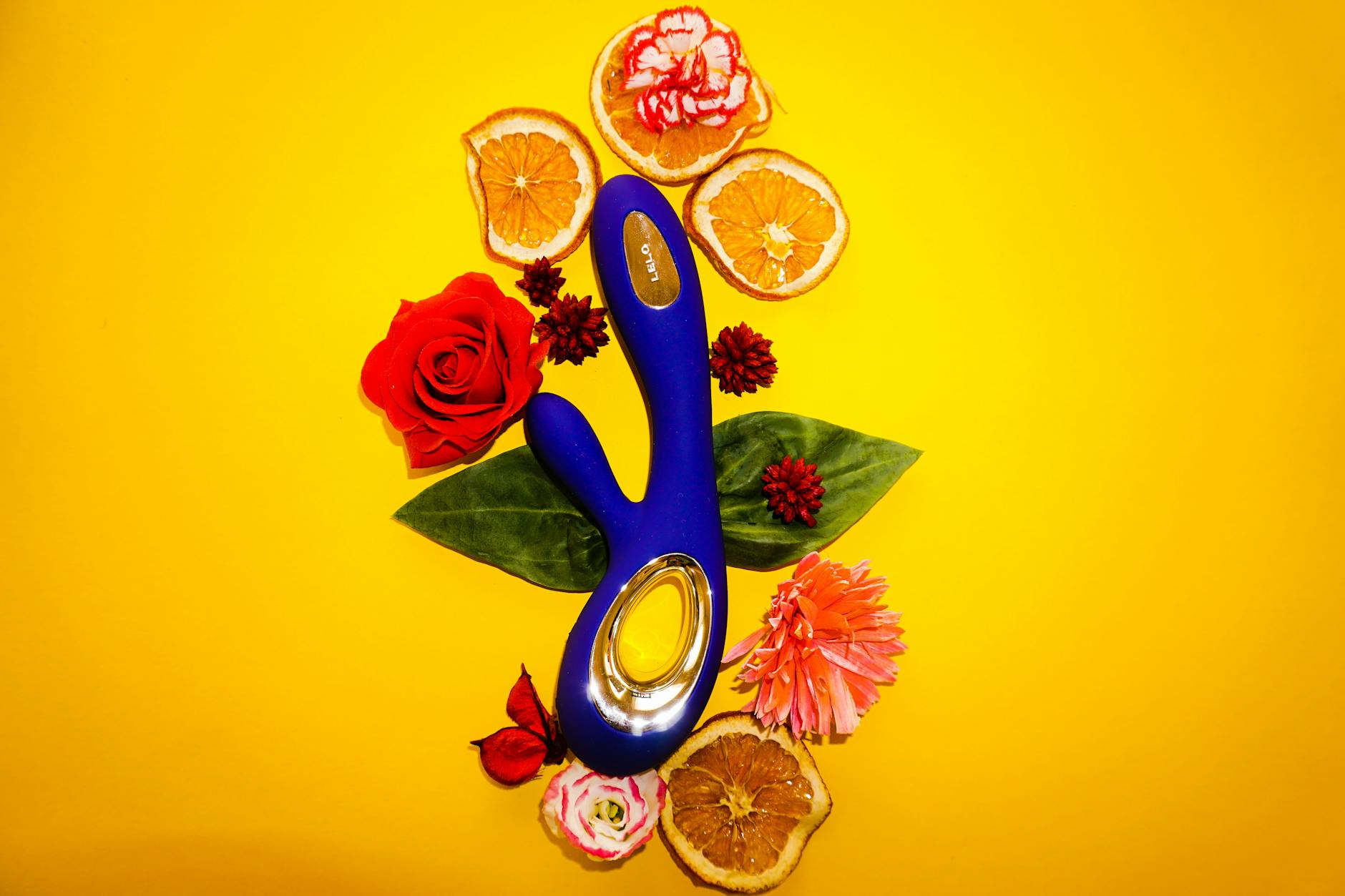 Blue lemon vibrator surrounded by flowers and citrus on a bright yellow background, symbolizing pleasure and intimacy.